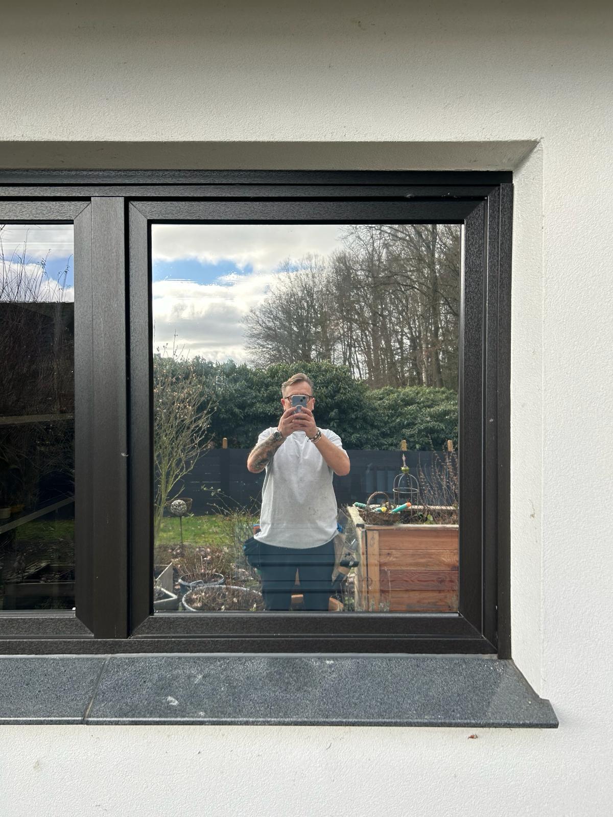 Fenster mit Spiegelung, klare Fensterfolie, dunkler Rahmen, Garten und Himmel sichtbar